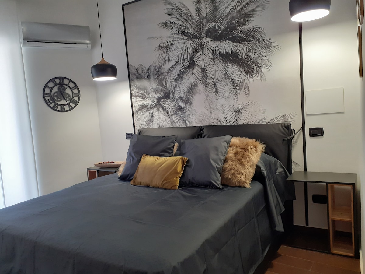 A spacious bed is dressed in gray linens, complemented by decorative pillows in neutral tones. A large mural featuring palm leaves creates a calming backdrop. Two pendant lights hang above the bed, adding soft illumination. A bedside table is positioned on one side.