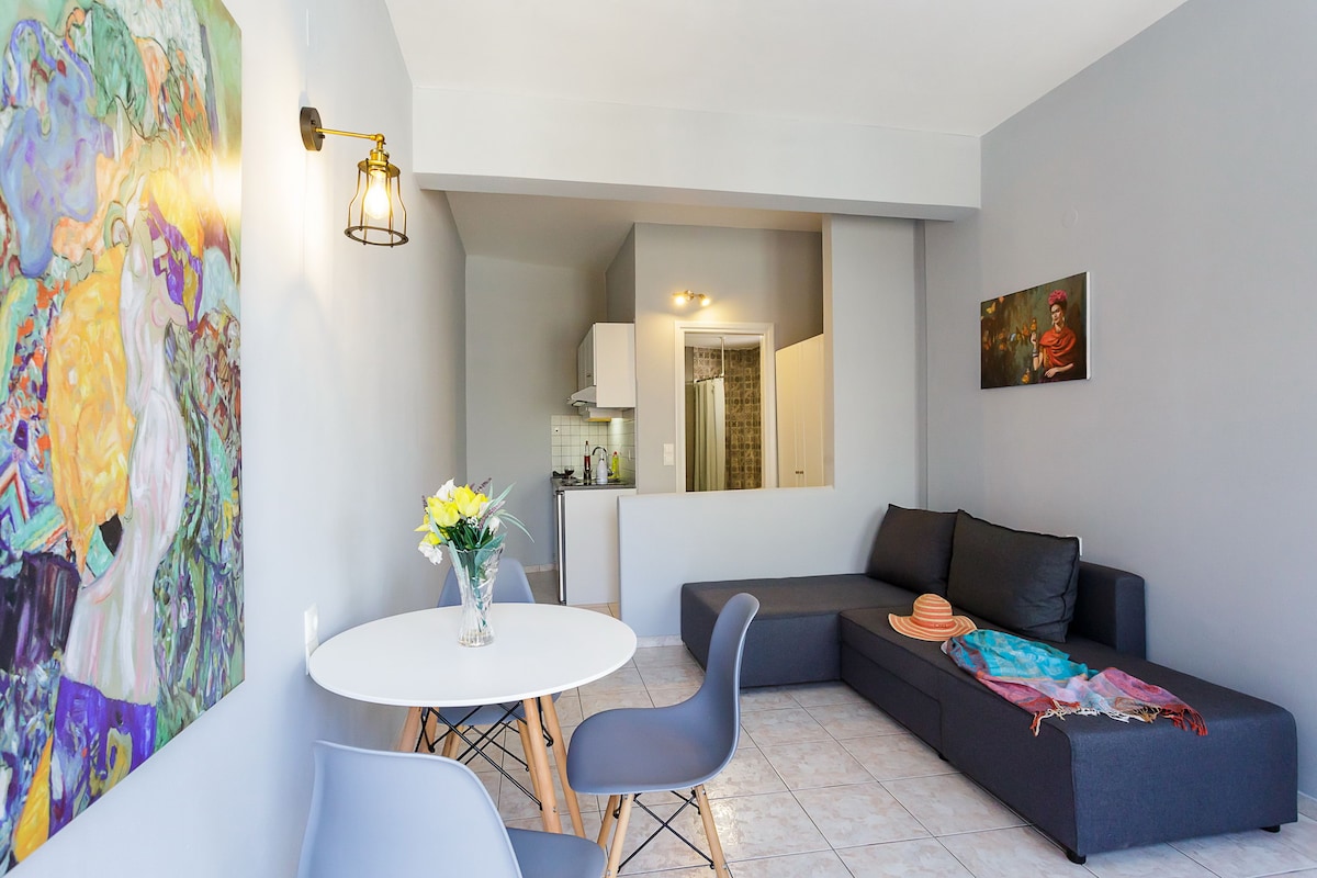 The open-plan living area features a comfortable sofa, a round dining table with chairs, and a kitchenette in the background. Light grey walls are adorned with vibrant artwork. A vase of flowers sits on the table, adding a touch of freshness to the space.