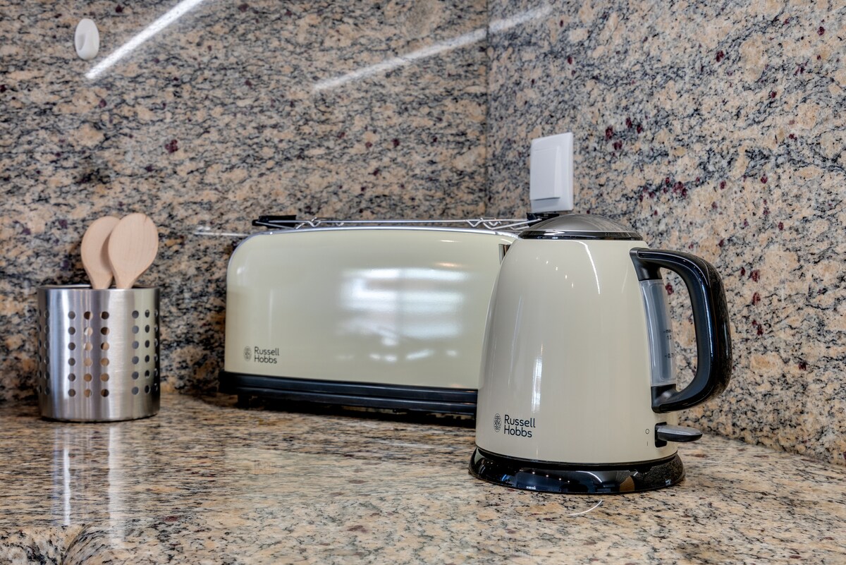 A modern kitchenette corner showcases sleek appliances on a speckled granite countertop. A stylish kettle and toaster are positioned beside a metallic utensil holder, with wooden cooking tools partially visible. The overall design emphasizes functionality and a clean aesthetic.