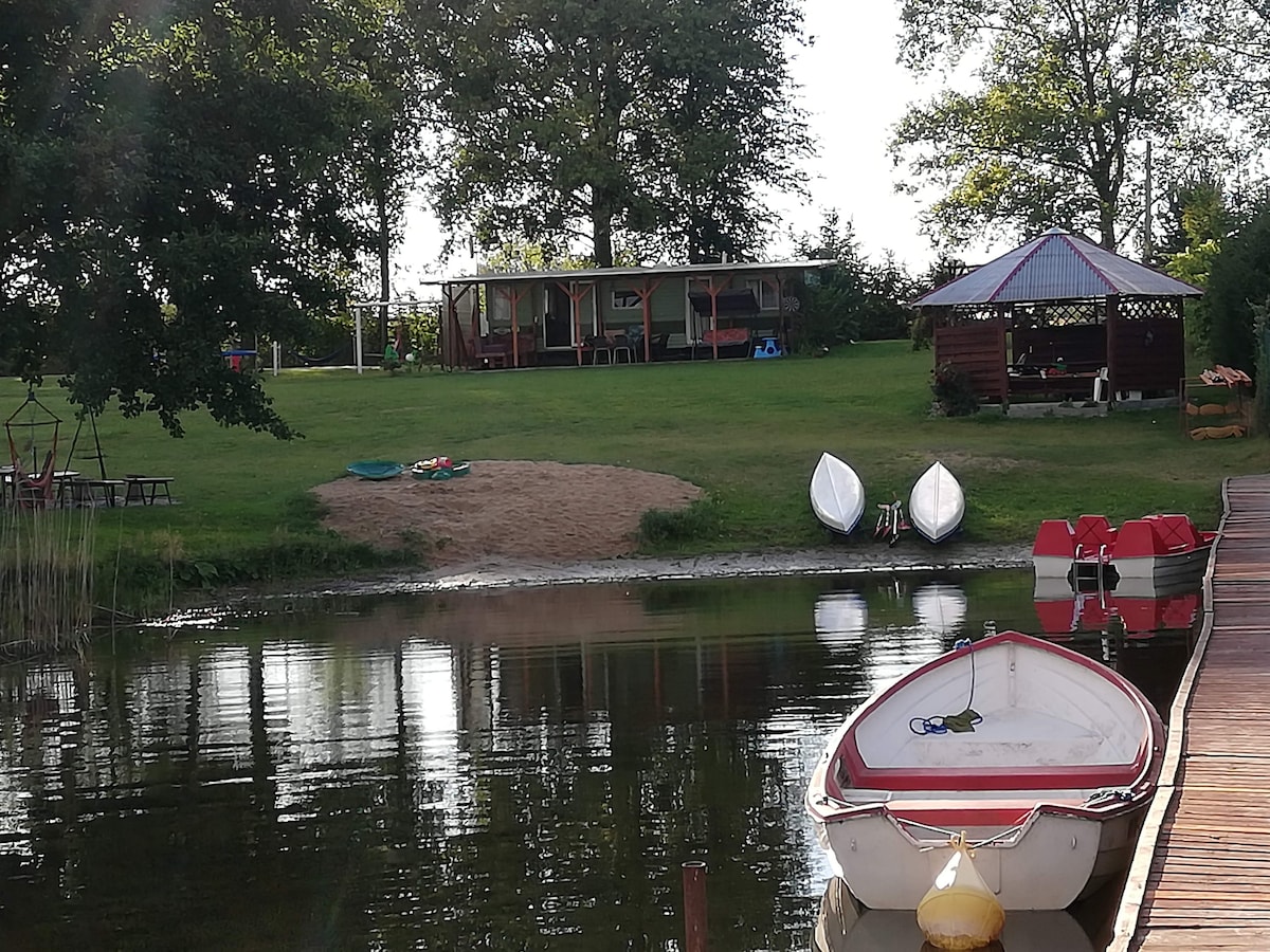 A calm lake scene features a white boat moored at the dock. In the background, a grassy area is visible with a small sandy beach and two lounge chairs. A gazebo is seen amidst trees, alongside several colorful watercraft resting on the shore.