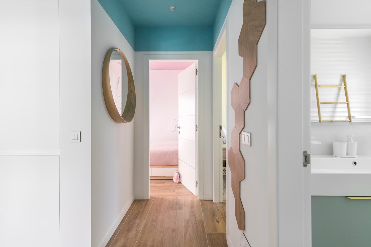 A bright hallway features a circular mirror and a textured wall art piece. Light wood flooring extends through the space, leading to an open door revealing a softly lit bedroom. The walls are painted in calming shades of white and turquoise.