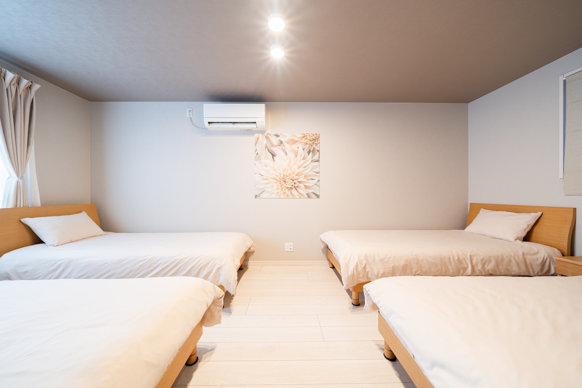 A bright sleeping area features four single beds arranged evenly, each with a light-colored bedding set. A floral wall art piece adds a touch of decor, while an air conditioning unit ensures a comfortable climate. Ample natural light enters through the window.