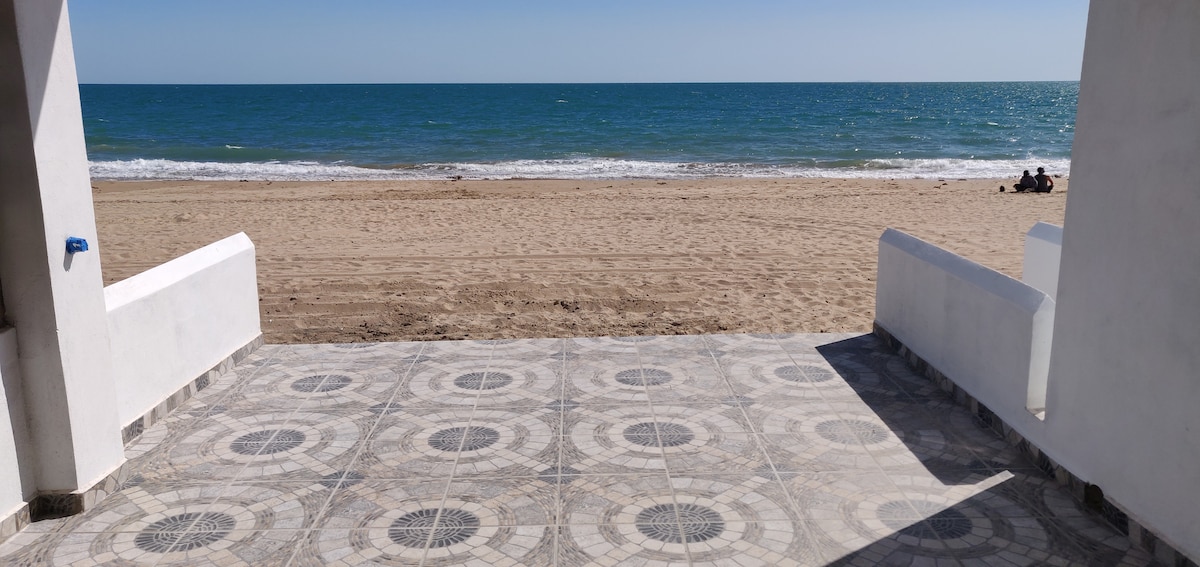 The view from a covered patio showcases sandy beachfront and the calm sea. The tiled floor exhibits a geometric pattern, complementing the serene coastal scenery. Gentle waves lap at the shore, inviting relaxation under clear blue skies.