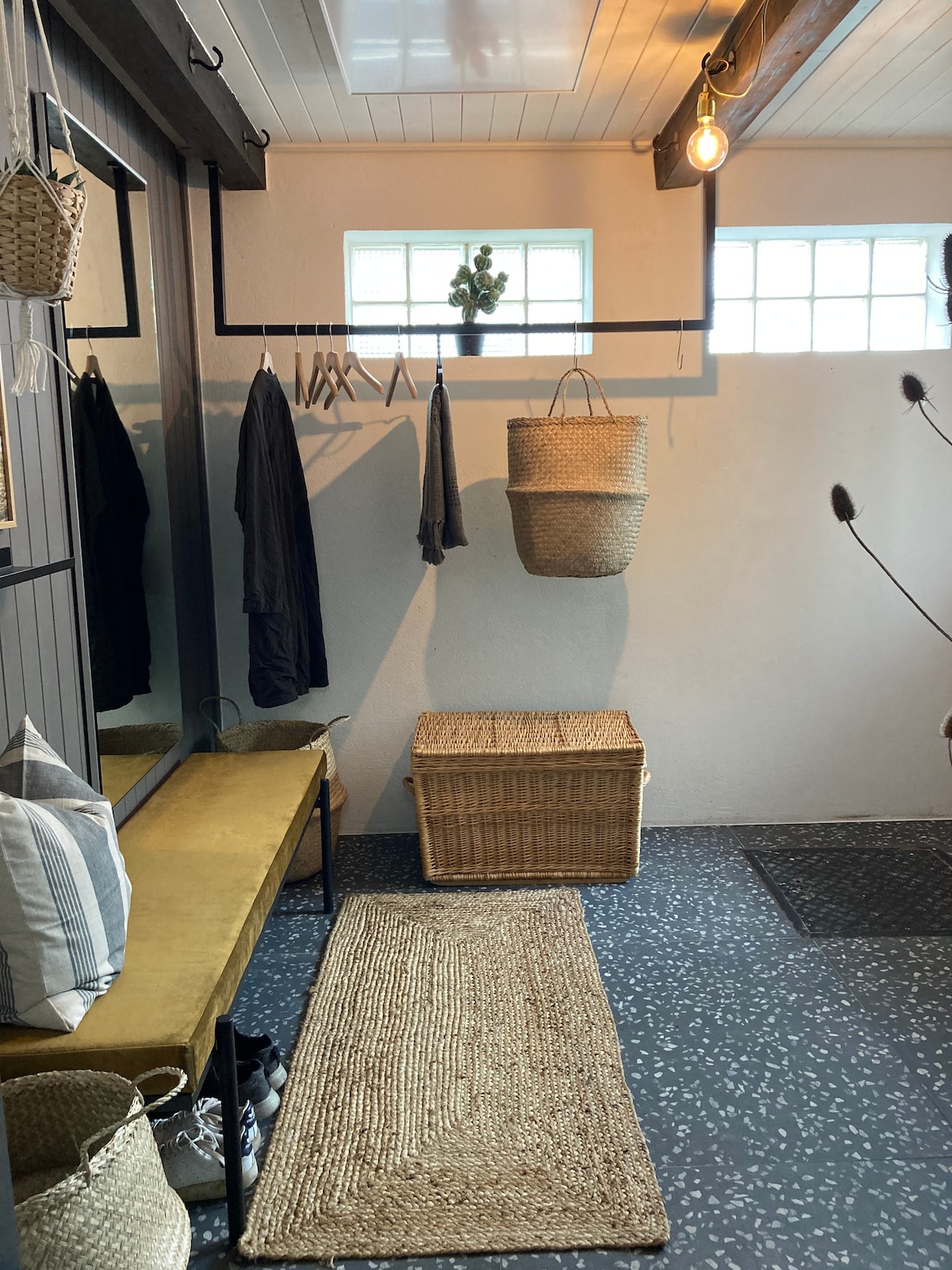 A welcoming entryway features a bench with a mustard-yellow cushion and a woven storage basket. Hooks line one wall, displaying clothing and accessories, while a textured rug leads to the door. Natural light filters through small windows, enhancing the inviting space.