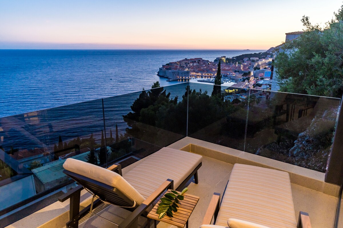 An outdoor terrace showcases two loungers, positioned to overlook the sea and the historical city center of Dubrovnik. The tranquil waters reflect soft hues from the twilight sky, while greenery frames the view, creating a peaceful atmosphere for relaxation.