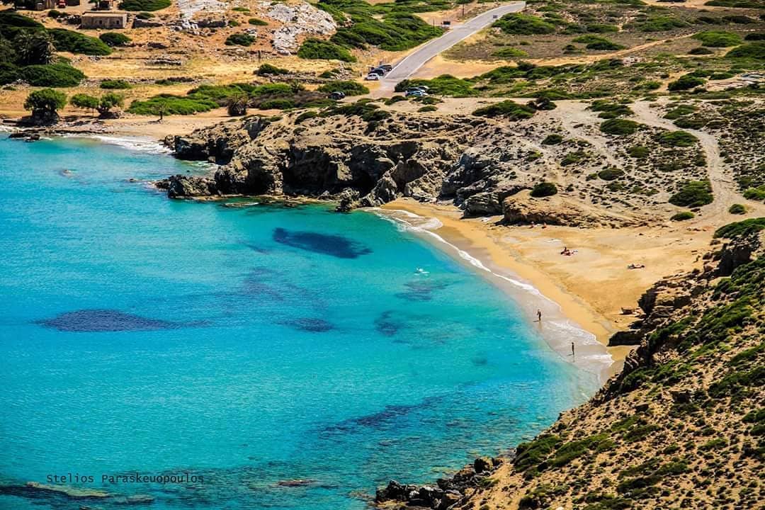 A tranquil beach scene features golden sand bordered by clear turquoise waters. Rocky cliffs rise along the shoreline. A few individuals can be seen enjoying the beach, while the surrounding greenery adds natural beauty to the landscape. A road runs parallel to the shoreline.