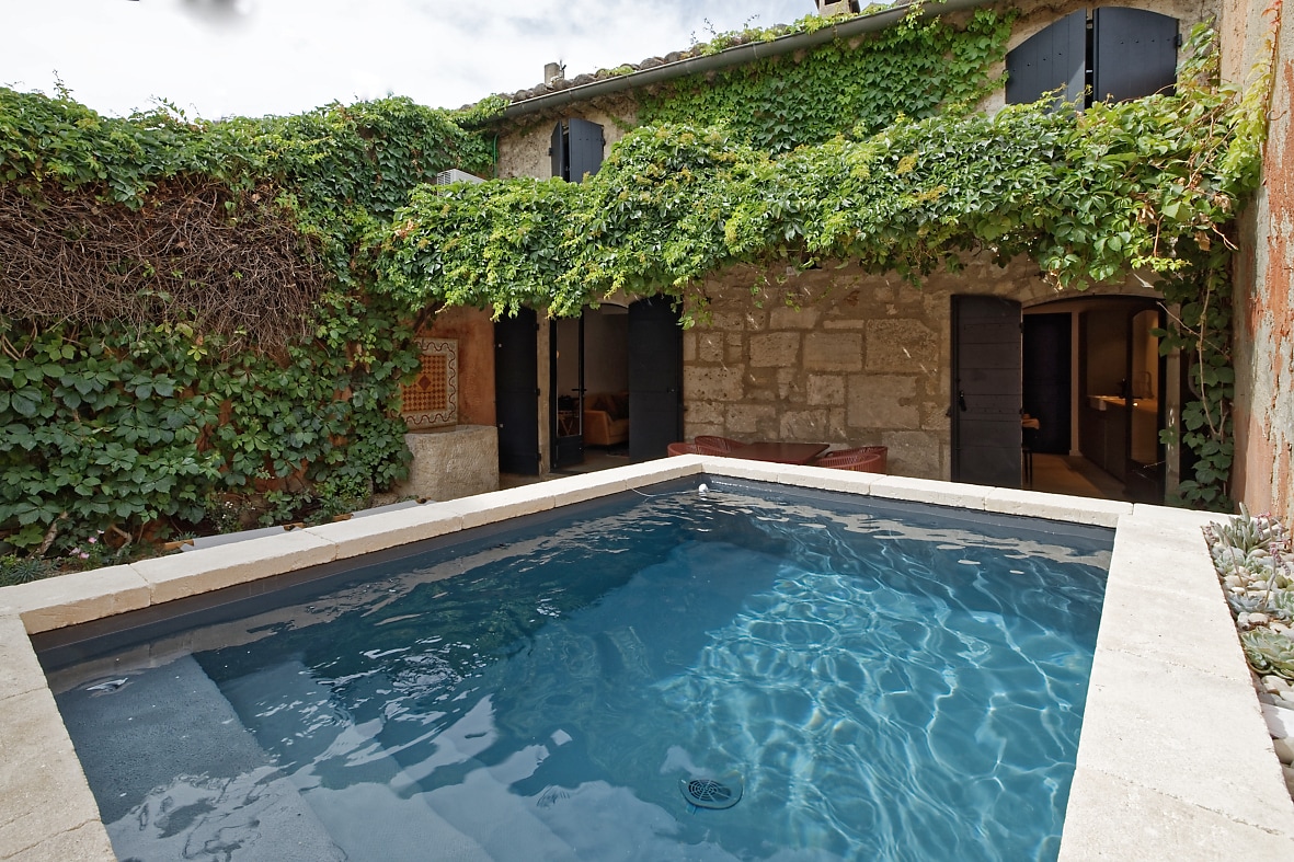 A private swimming pool is surrounded by lush greenery, with stone walls and dark wooden shutters in the background. The clear water reflects the sky and is framed by a tiled edge, creating an inviting outdoor space.