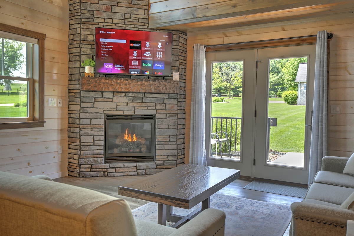A cozy living area showcases a stone fireplace beneath a flat-screen TV, with soft lighting accenting the wooden beams overhead. Large doors provide access to the outside, revealing a grassy landscape. A neutral-toned sofa is paired with a rustic coffee table, inviting relaxation.