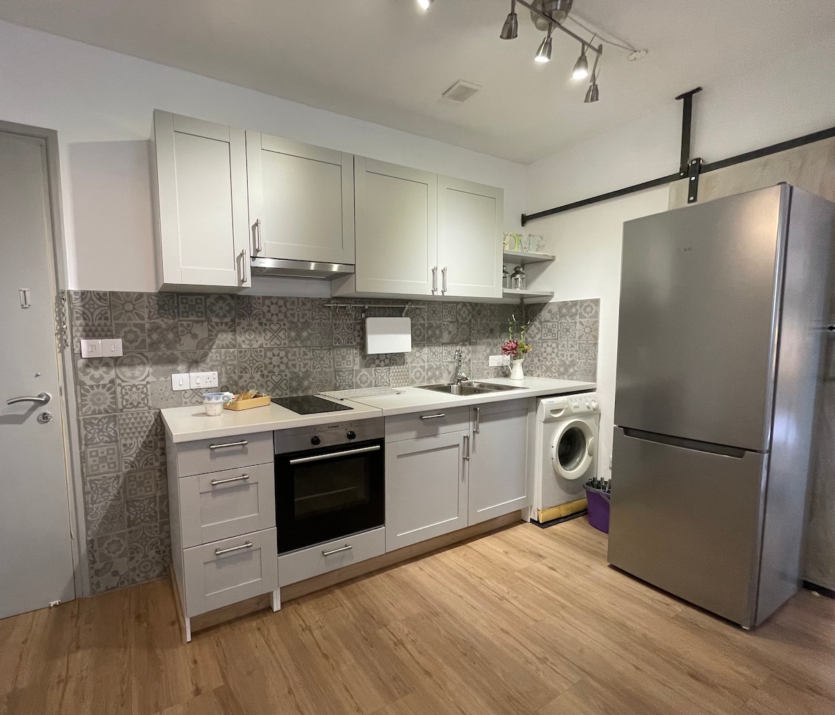 A modern kitchen with light grey cabinetry and stainless steel appliances is showcased. The patterned backsplash adds visual interest. An oven, cooktop, and refrigerator are distinctly visible. A washing machine is positioned in the corner, with wooden flooring completing the warm, functional space.