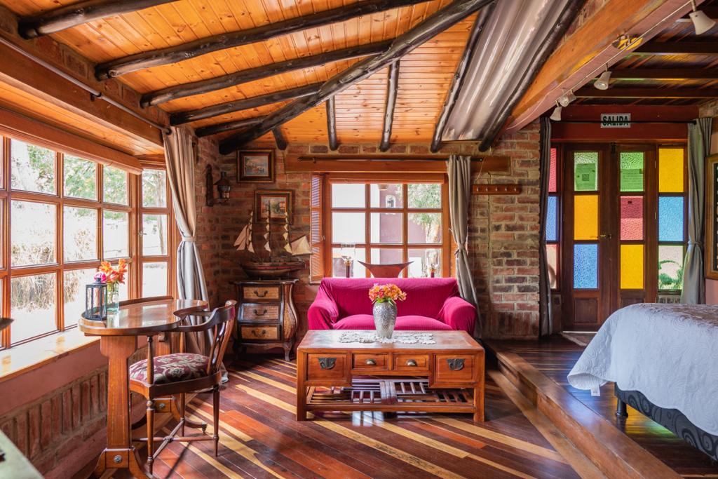 A bright living area features a pink sofa centered across polished wooden floors. A small dining table is set near a window, complemented by fresh flowers. Colorful stained glass accents are visible in the entrance door, enhancing the room's natural light and inviting ambience.