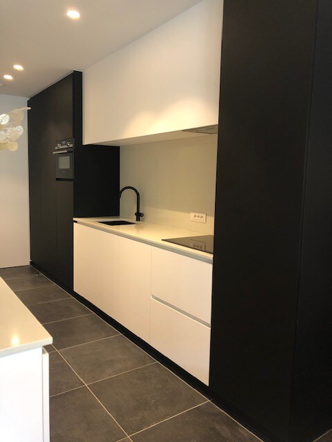 A modern kitchen features sleek white cabinetry combined with dark accents. A minimalist countertop is complemented by a built-in stovetop and sink with a matte black faucet. Ambient lighting enhances the contemporary design, while the floor is covered in dark tiles.
