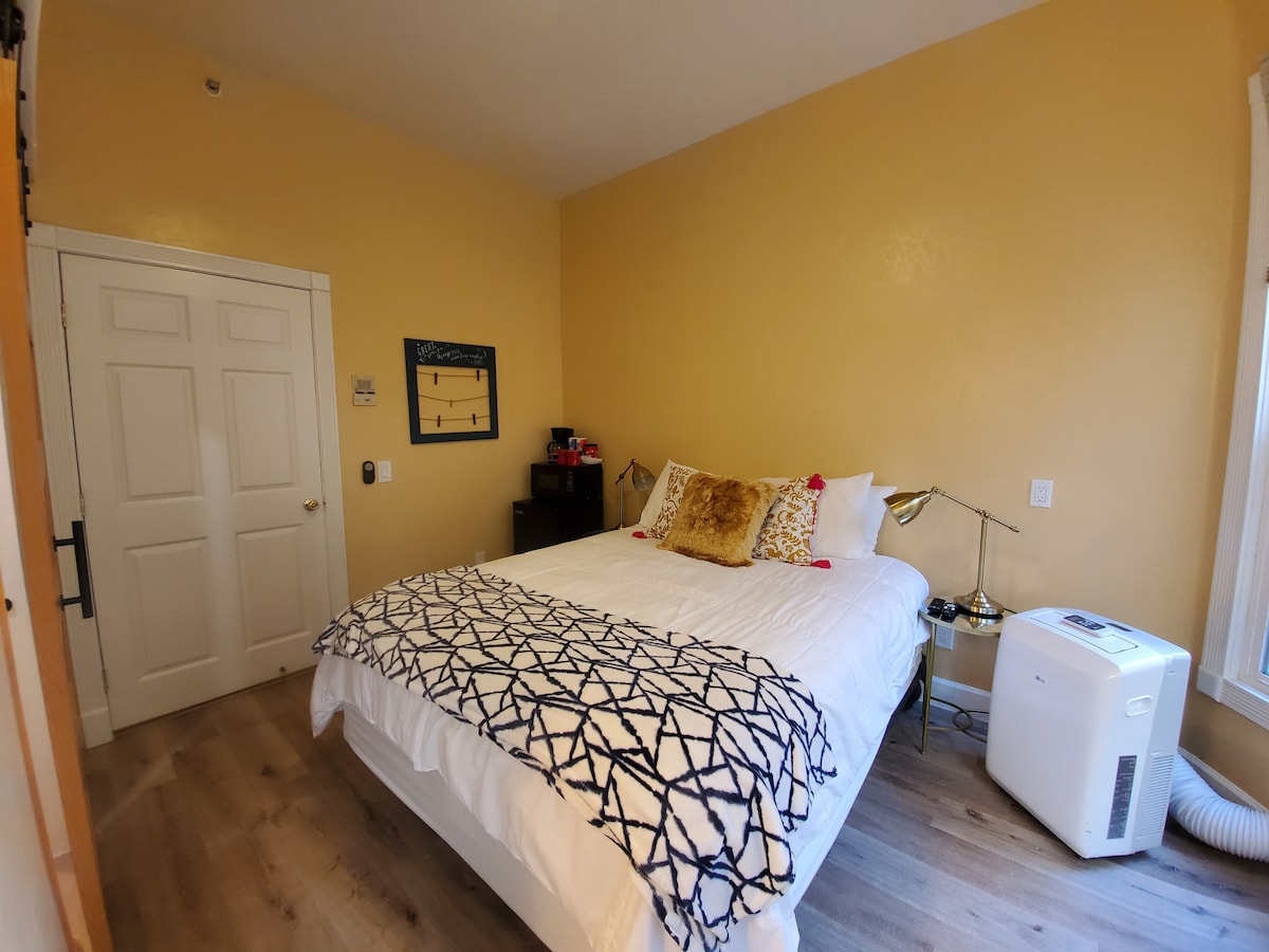 A well-appointed hotel room features a spacious bed dressed in white linens with decorative pillows. A simple geometric throw blanket is positioned atop the bed. The walls are painted a warm yellow, and a lamp stands next to the bed, providing additional lighting.