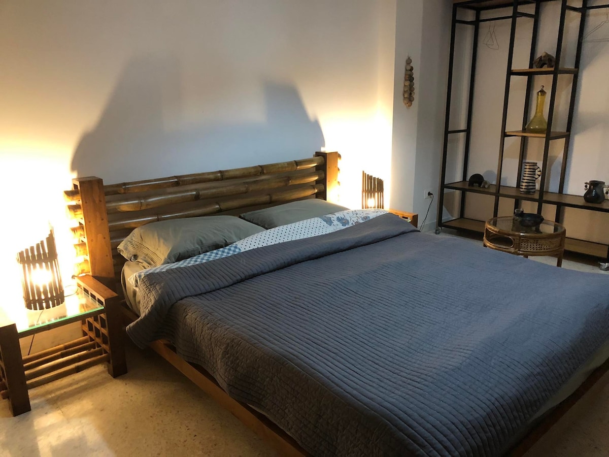 A spacious sleeping area features a large bed with neutral-colored bedding. Soft lighting is provided by lamps on either side, casting a gentle glow. A minimalist shelving unit displays decorative items, contributing to the calming environment of the room.