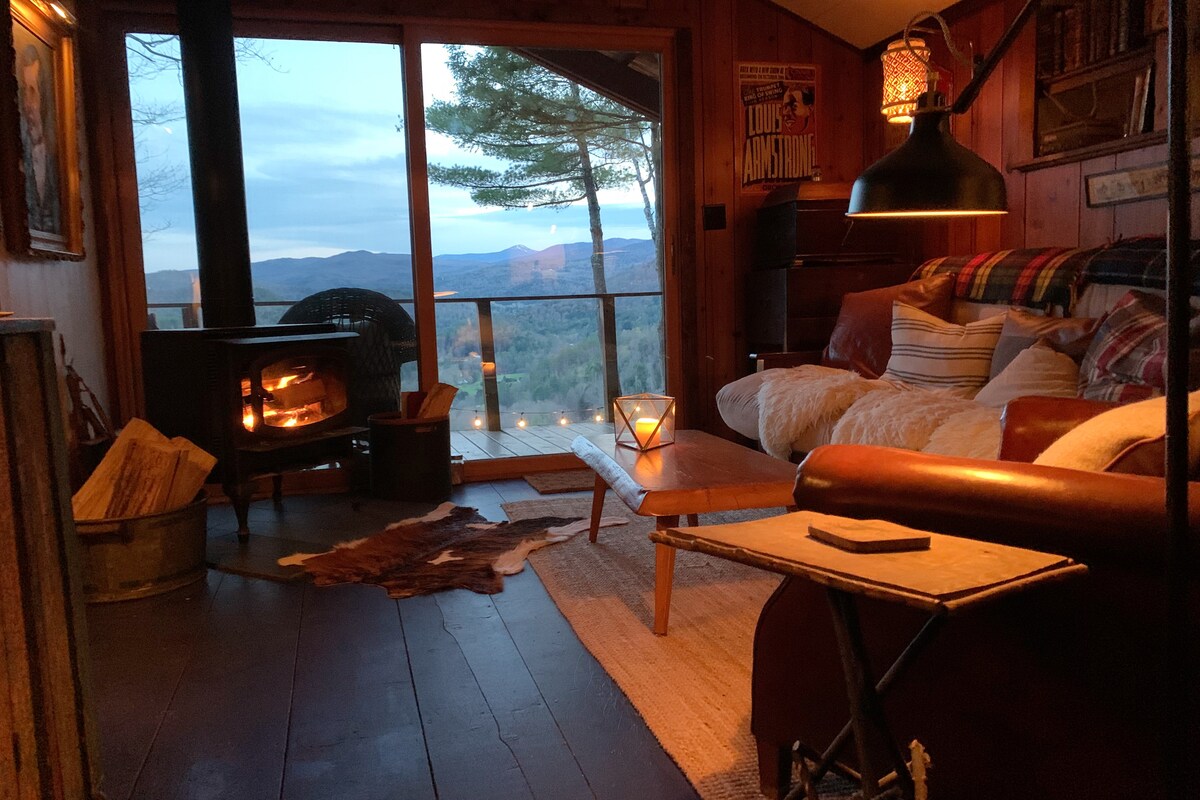 A cozy sitting area is illuminated by a warm glow from a wood-burning stove. Vintage furnishings, including a leather sofa and a rustic coffee table, complement large glass doors that frame expansive mountain views in the distance. Natural light filters softly through the room.