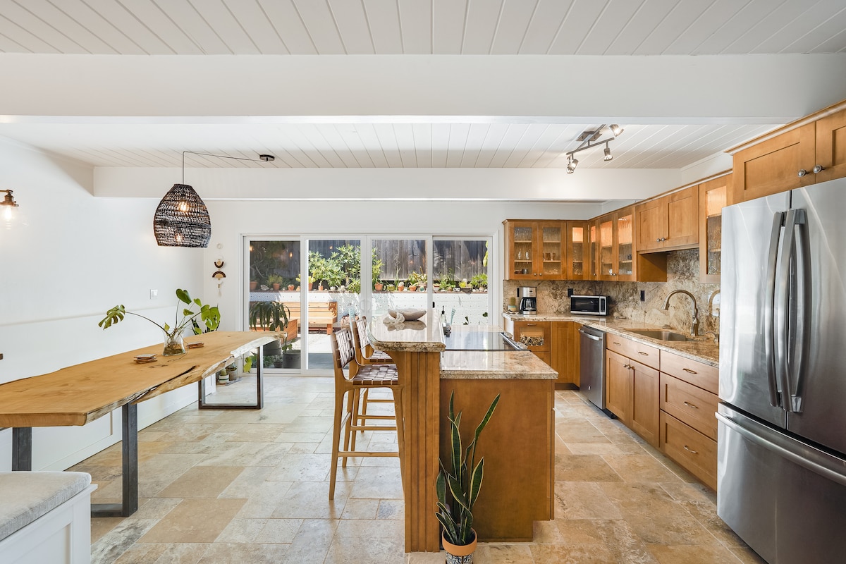 A spacious kitchen features a large island with seating and wooden cabinetry. Natural light fills the area through sliding glass doors that lead to a garden. A dining table made of wood complements the modern appliances and finishes, creating an inviting space for meal preparation.
