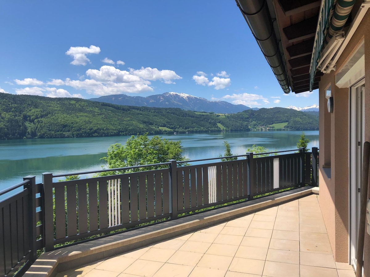 A scenic terrace overlooks a lake, with lush green hills and distant snow-capped mountains framed against a clear blue sky. The area features a tiled floor and a railing, providing an inviting outdoor space for relaxation.