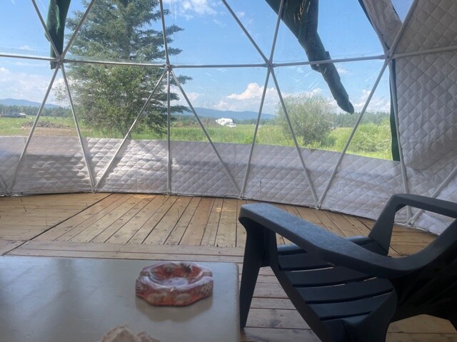The interior of the geodesic dome features wooden flooring and a spacious seating area. Large windows provide panoramic views of the surrounding landscape and distant mountains. A black chair and a small table are positioned next to a decorative object, allowing for relaxation and enjoyment of the outdoors.