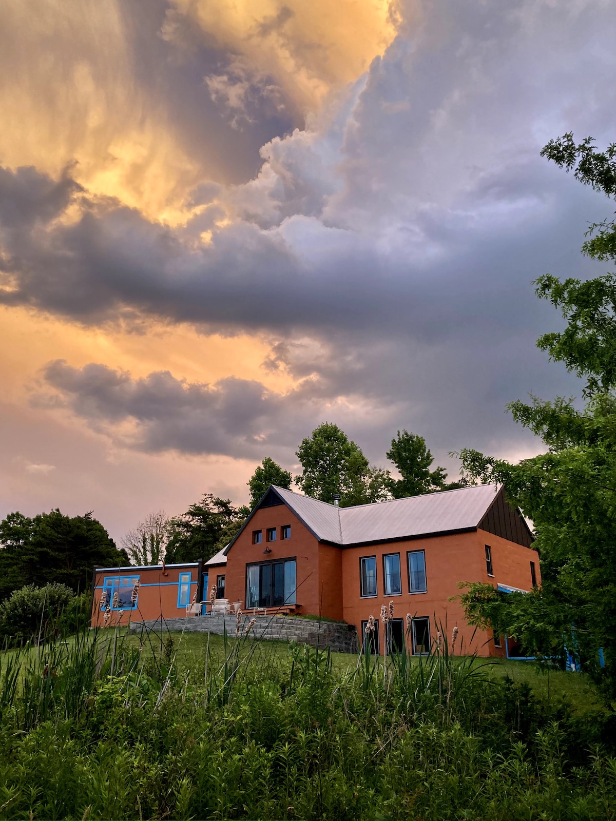 A modern home features warm orange exterior walls and a pitched roof, set against a dramatic sky filled with yellow and gray clouds. The property is surrounded by lush greenery, enhancing the tranquil country atmosphere.
