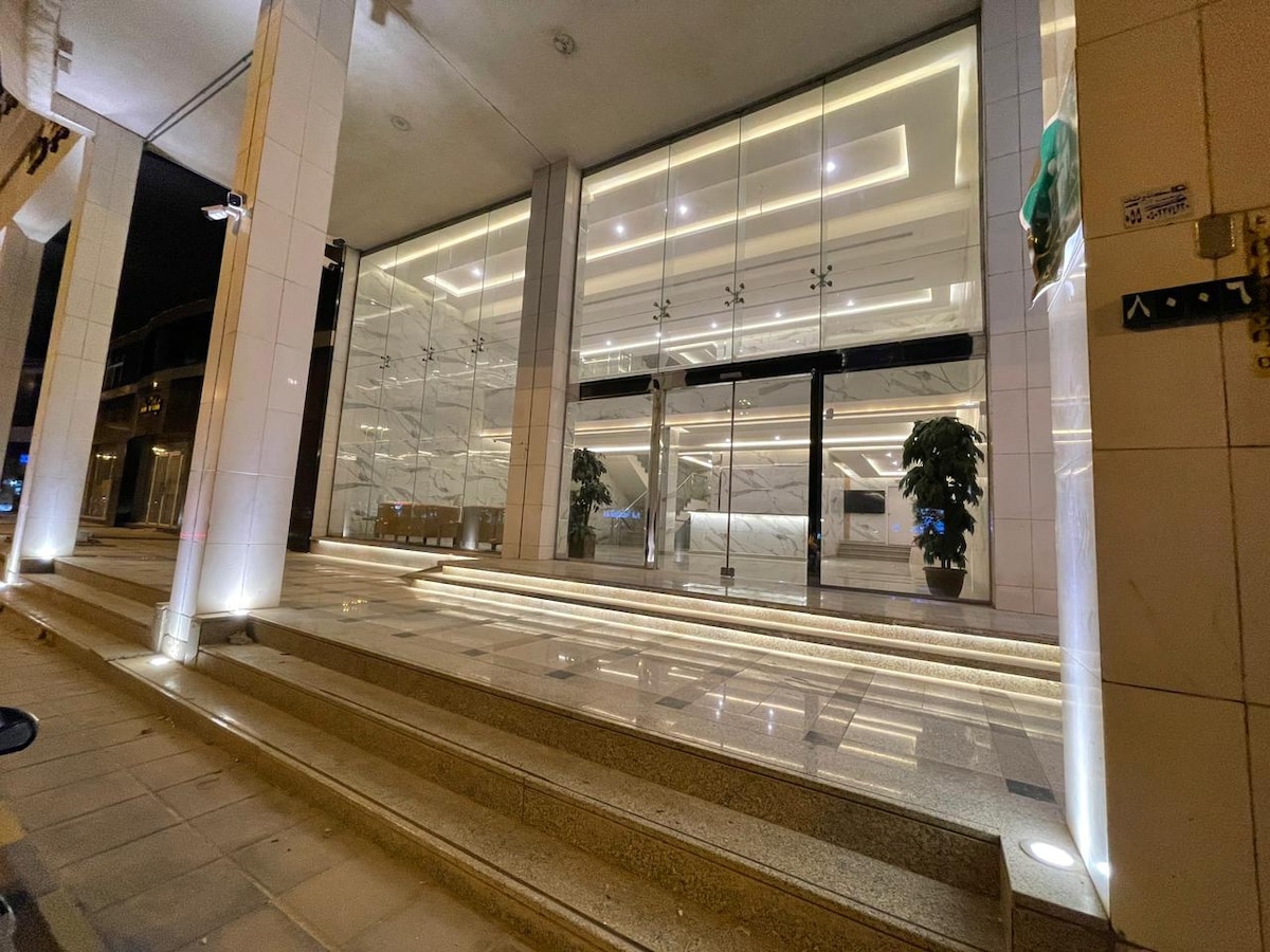 The entrance features a modern facade with large glass doors and sleek marble steps. Soft lighting highlights the architectural details, creating an inviting atmosphere. Potted greenery flanks the entrance, enhancing the urban setting.
