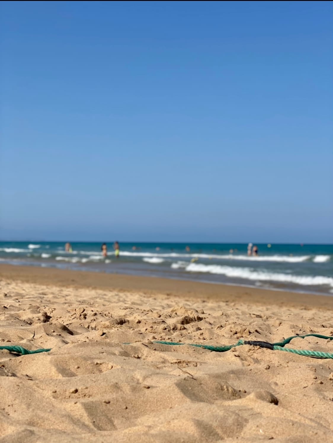 A sandy beach stretches out under a clear blue sky, with gentle waves lapping at the shore. Figures can be seen enjoying the water, while the distant horizon meets the calm sea, creating a serene coastal atmosphere.