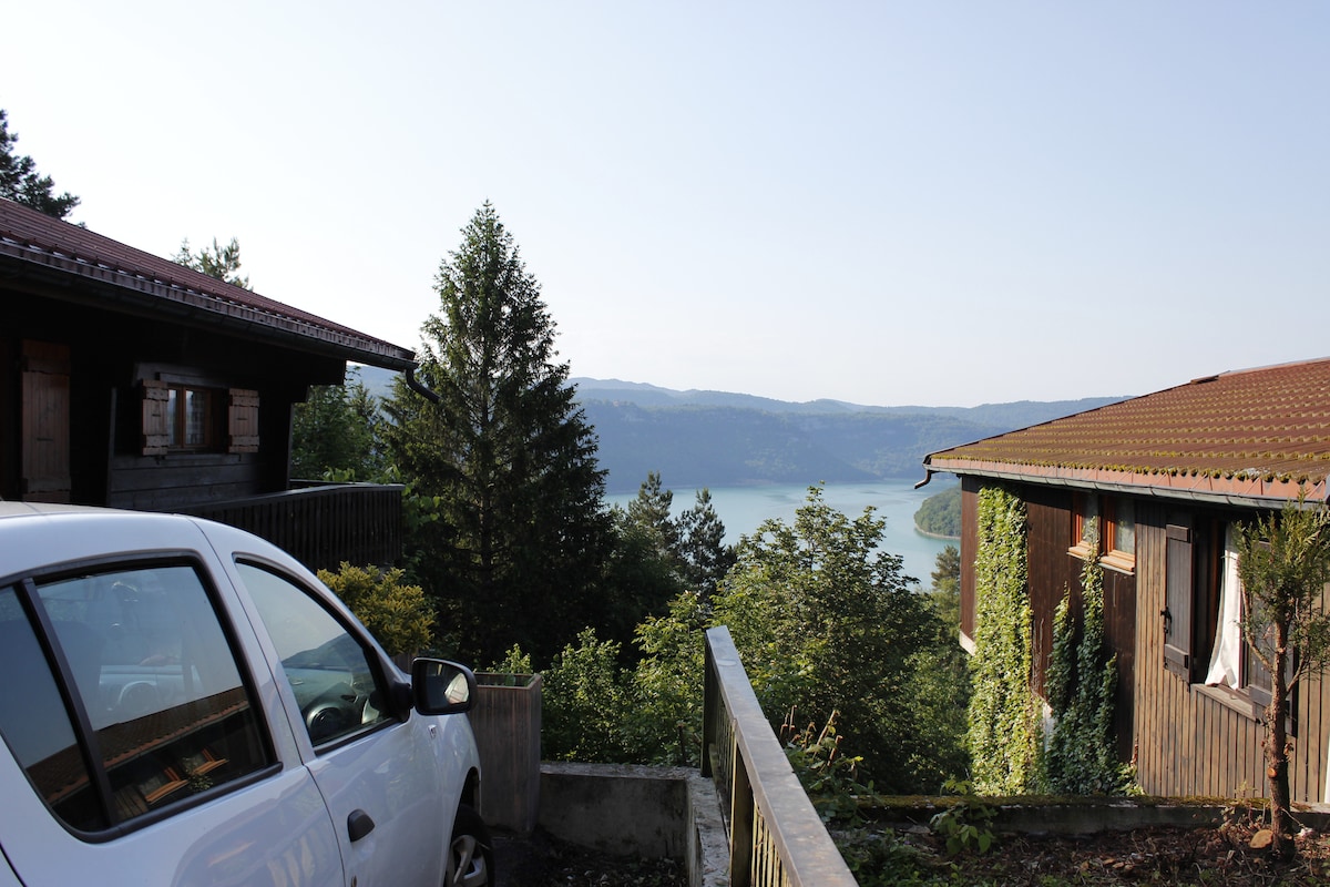 A view reveals two wooden chalets in a serene landscape, with a car parked nearby. Lush greenery surrounds the area, leading towards a lake visible in the background, framed by distant hills under a clear sky.