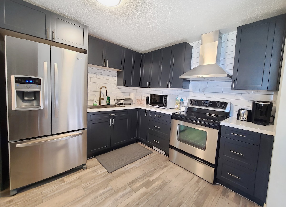 The modern kitchen features sleek dark cabinetry and stainless-steel appliances, including a refrigerator, oven, and microwave. A white tile backsplash complements the space, while ample counter space hosts a coffee maker and small kitchen appliances. A light wood floor adds warmth to the contemporary design.