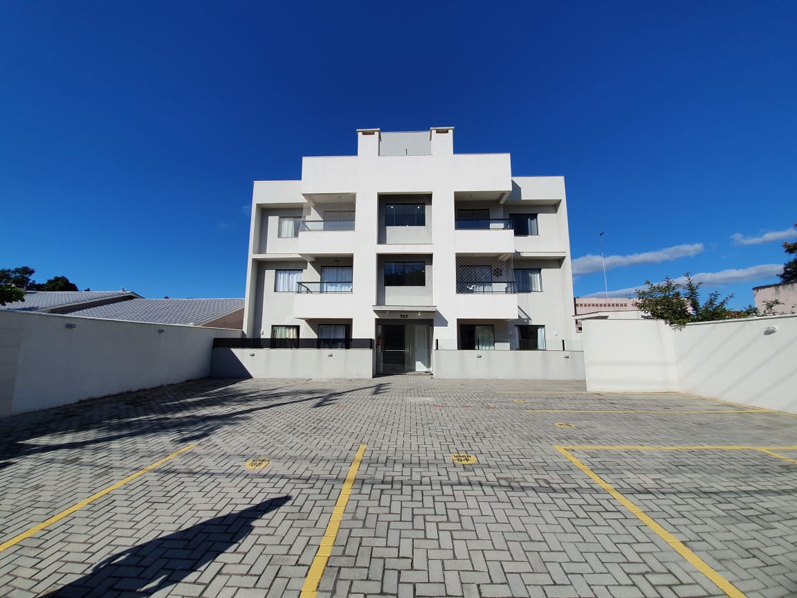 A three-story building features a modern architectural design with a clean white exterior. The spacious parking area includes designated spaces marked on the paved surface, with a clear sky visible above. Large windows adorn the front, allowing natural light to enter.