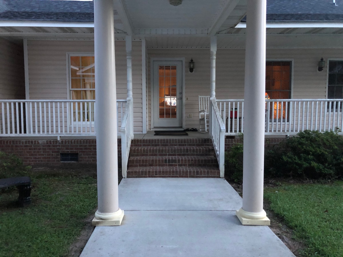 The welcoming entrance features a concrete pathway leading to a porch supported by white columns. Two sets of steps provide access to the front door, which is framed by warm light from the interior. Lush greenery is present along the sides.