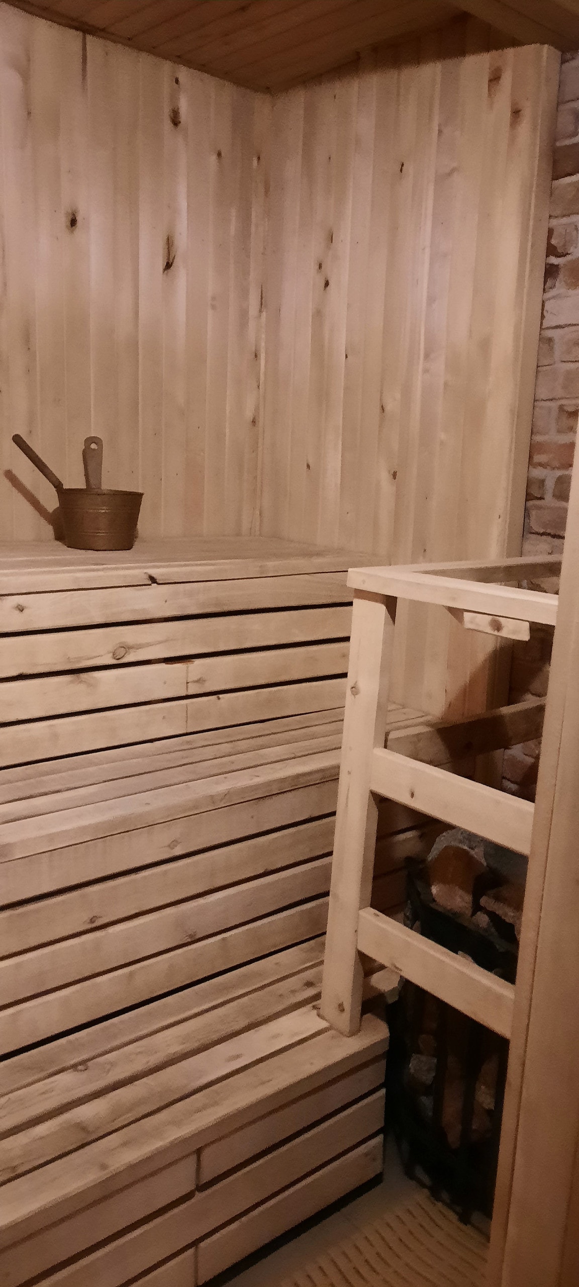 A wooden sauna interior features smooth planks and benches, creating a warm atmosphere. A metal bucket and ladle are positioned on a bench, while a storage shelf displays neatly arranged wood logs, complementing the natural wood finish of the space.