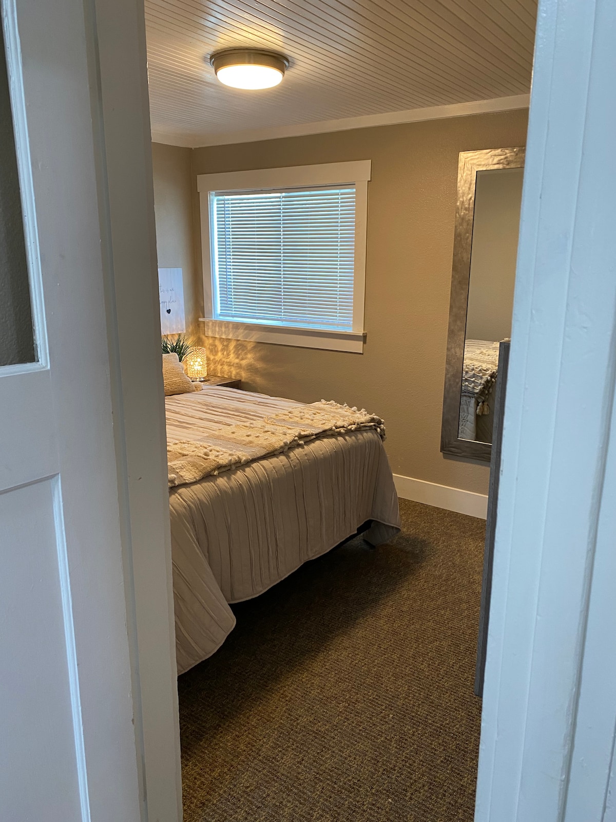A well-lit bedroom is accessed through an open door, featuring a spacious bed with a textured beige coverlet. Natural light enters through a window with white blinds, and a decorative mirror is positioned nearby, enhancing the room's bright and airy feel.