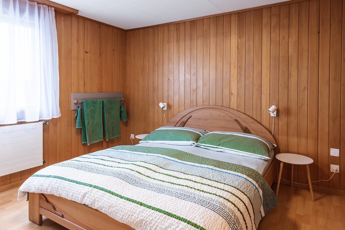A warm wooden paneling surrounds a cozy sleeping area. A spacious bed is dressed in a white and green patterned blanket. Two reading lights are mounted on the wall, and a small side table is positioned beside the bed. Fresh towels hang neatly on a rack.