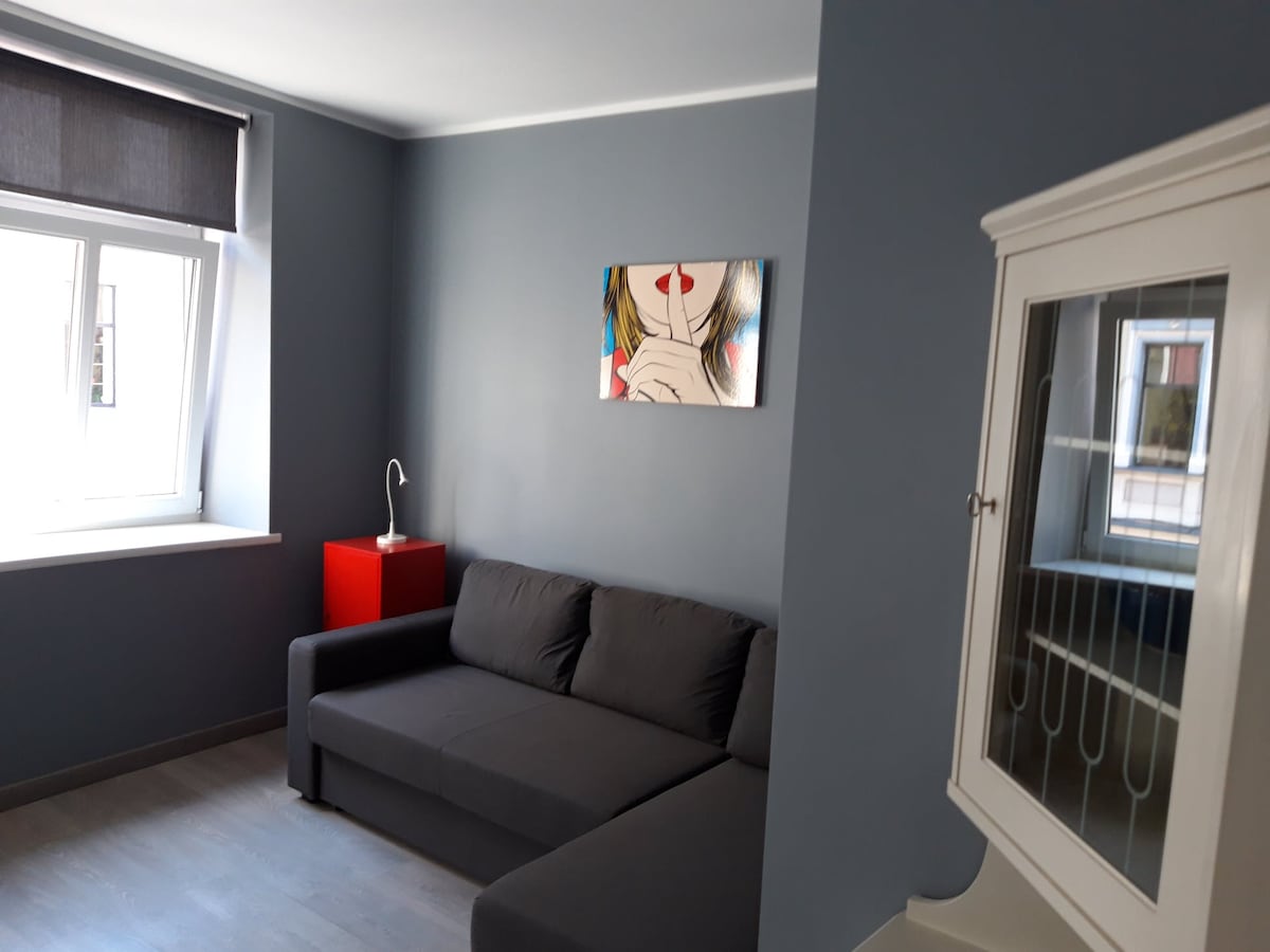 A modern living area is highlighted by a dark gray sofa positioned against a softly painted blue wall. Natural light enters through a large window with a roller shade, and a decorative artwork featuring a woman's face adds visual interest. A small red side table stands adjacent to the sofa.