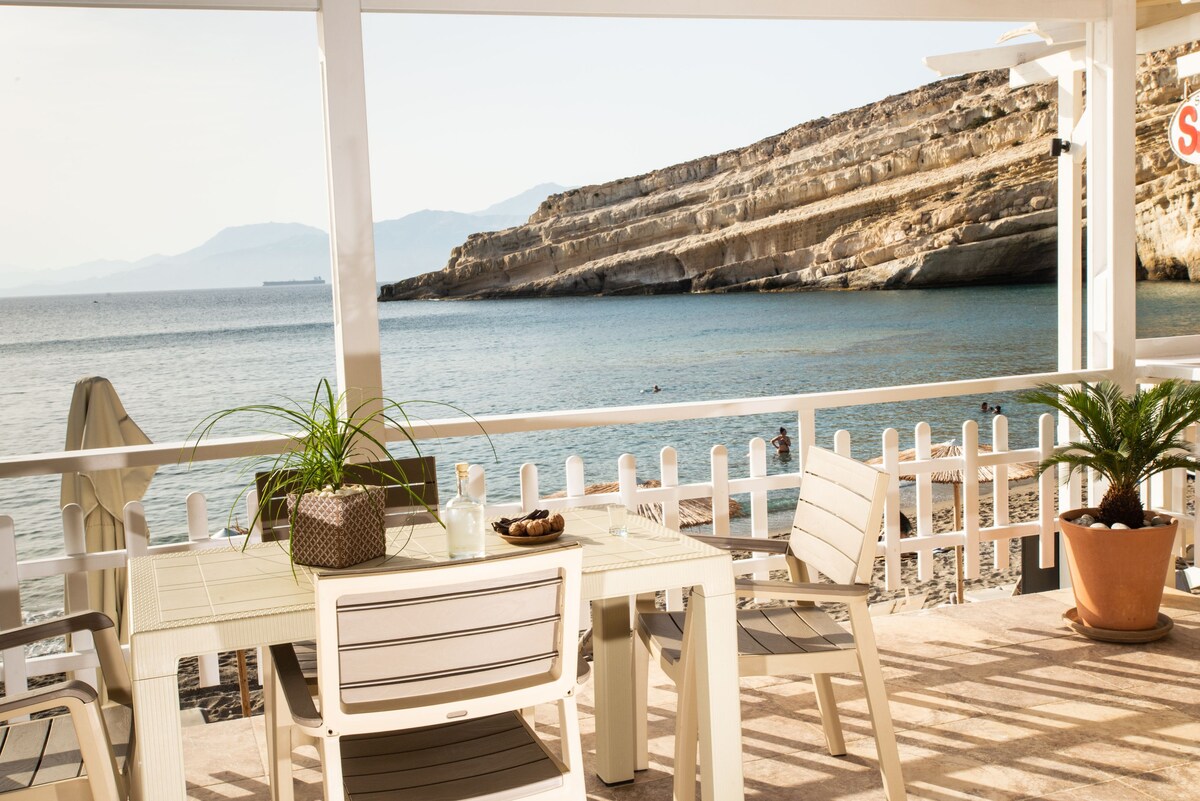A spacious terrace is presented, featuring a table and several chairs. Potted plants add natural greenery. The view overlooks the tranquil sea and distant cliffs, while the beach is visible through the railing. Soft sunlight casts a warm glow across the outdoor seating area.