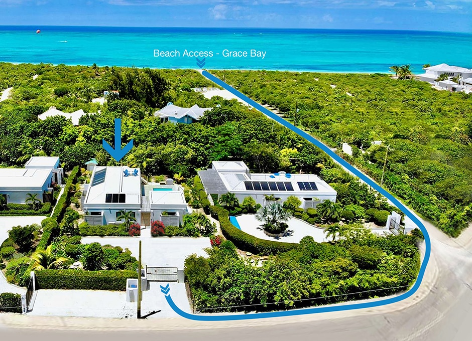 An aerial view of Beach Please Villa is shown, highlighting its proximity to Grace Bay Beach. The villa is surrounded by lush greenery, with a clear pathway leading to the beach access. The vibrant blue ocean is visible in the distance, enhancing the tranquil setting.