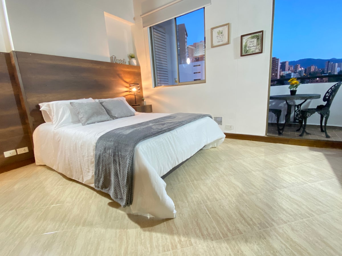 A spacious sleeping area is furnished with a cozy bed, featuring neutral bedding and decorative pillows. A balcony is visible through a large window, providing views of the city skyline. A small dining set is positioned on the balcony, enhancing the room's charm.
