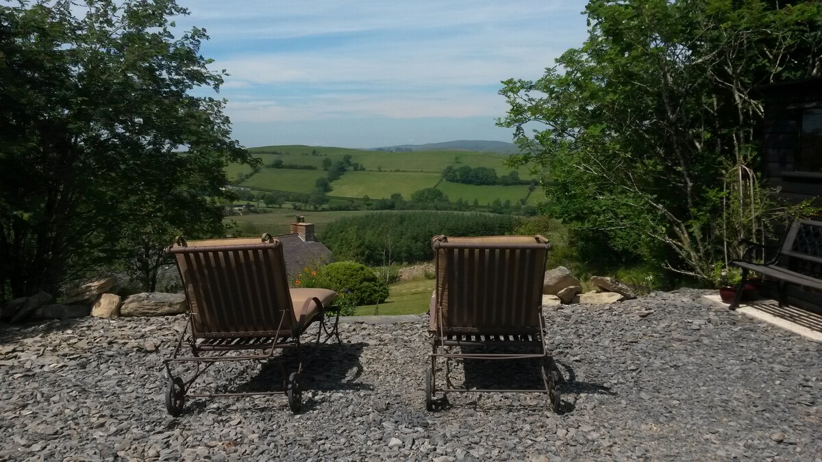 Two reclining chairs are positioned on a gravel area, facing expansive green hills and trees. The serene landscape is visible under a clear sky, with distant mountains providing a picturesque backdrop.