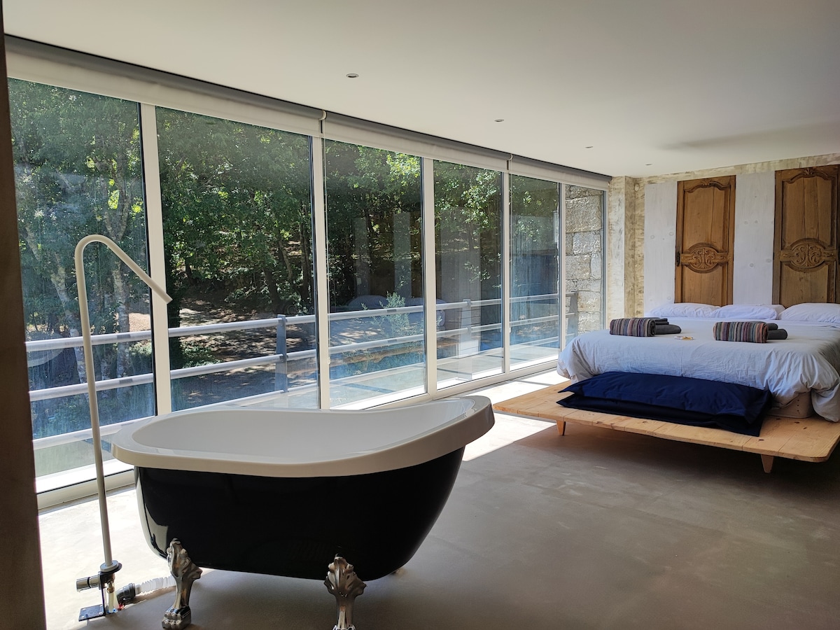 A stylish bedroom showcases a spacious bed with neatly arranged linens, positioned near large floor-to-ceiling windows that invite natural light. A classic black freestanding bathtub adds a touch of elegance, while the surrounding forest views enhance the serene environment.