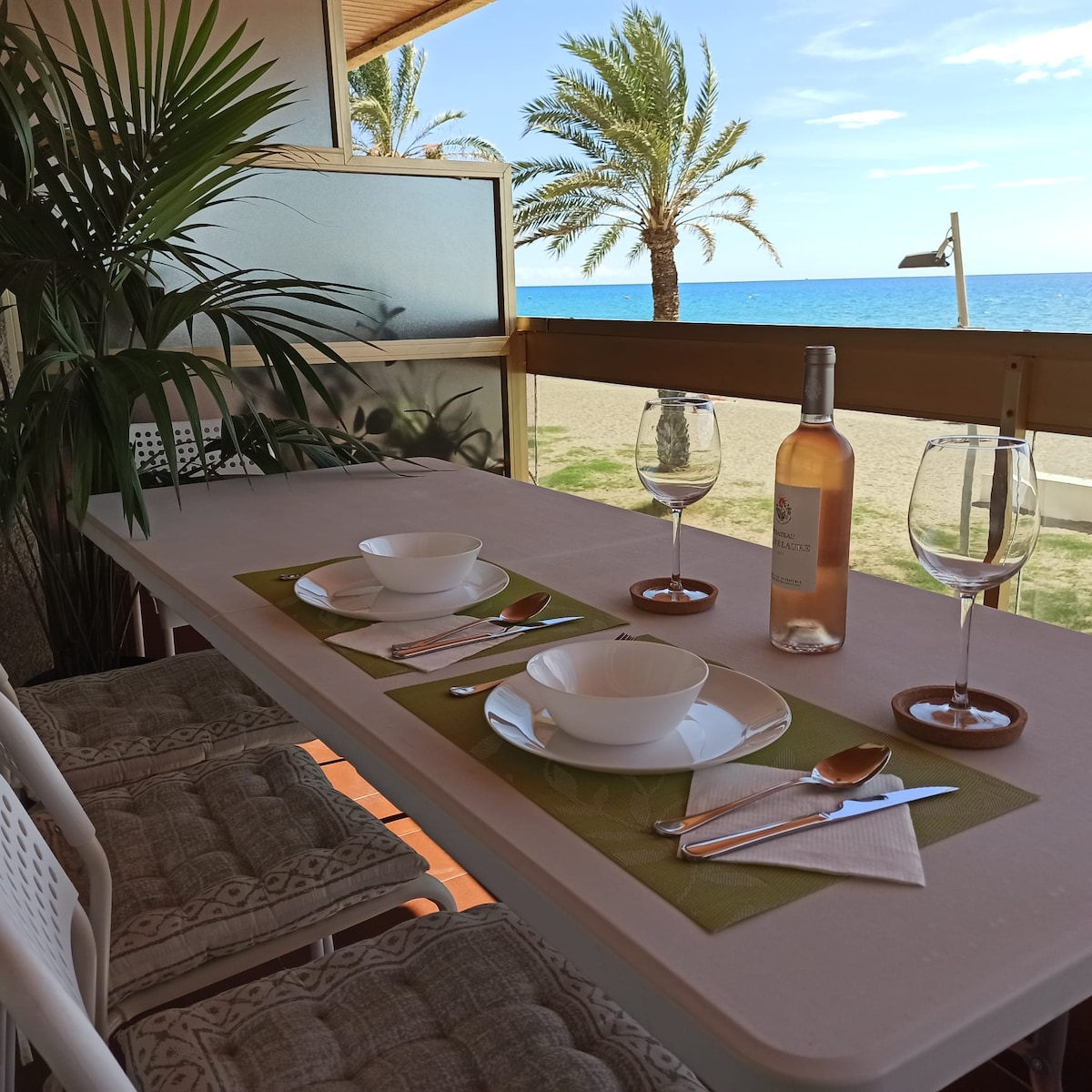 A dining table is set for two, featuring plates, glassware, and utensils on a green placemat. The backdrop showcases a beach view with palm trees and blue sea, enhancing the outdoor dining experience.