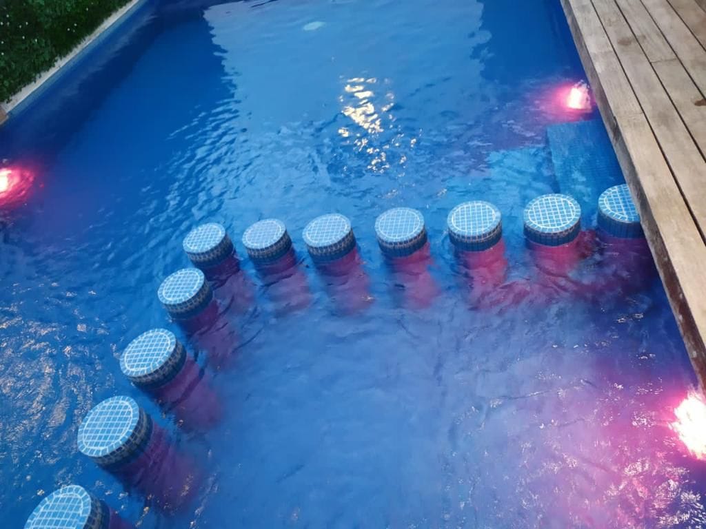 A swimming pool featuring decorative seating areas partially submerged, arranged in a circular pattern. The pool is illuminated with soft lighting, creating a calming ambiance. The water surface reflects the surrounding landscape and the colors from the lights.