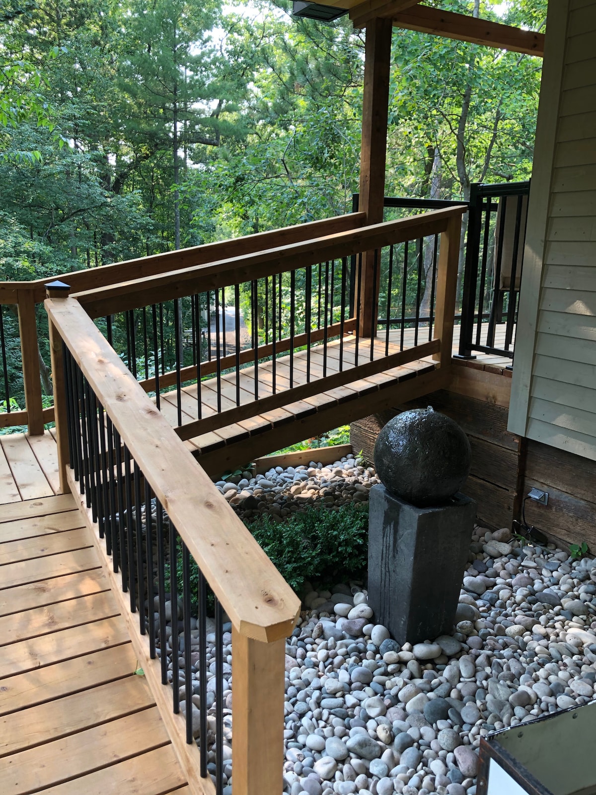 A wooden staircase with a metal railing leads upward through a serene outdoor setting. River stones are arranged around a stone pedestal featuring a spherical sculpture. Lush greenery frames the scene, contributing to a peaceful and natural ambiance.