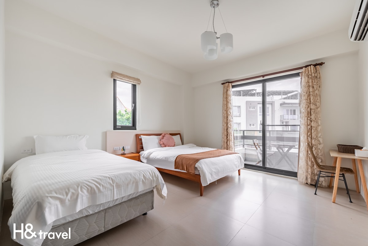 A bright and airy room features two beds, one with a chocolate brown throw blanket. Large windows allow natural light to fill the space. A simple wooden desk is positioned against a wall, and light curtains frame the windows for a modern touch.