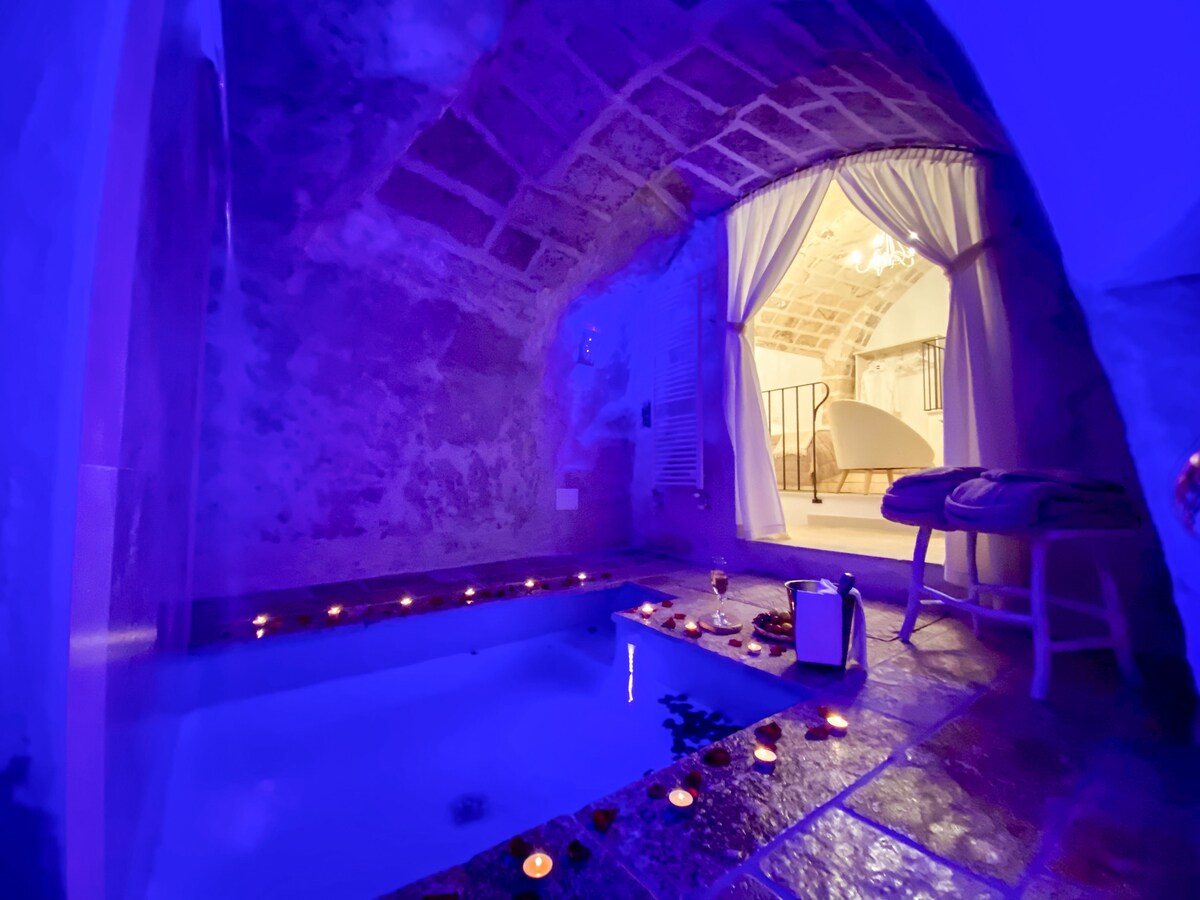 A serene spa area is nestled within a natural cave, illuminated by soft blue lights. Candles line the edges of a warm bath filled with water, creating a calming ambiance. A doorway leads to a spacious bedroom area, partially obscured by sheer curtains.