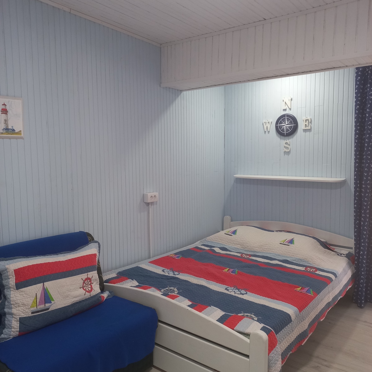 A cozy sleeping area features a bed adorned with a patterned quilt in blue and white hues. A small accent chair sits beside the bed, complementing the nautical-themed decor, while a wall clock and decorative elements add a serene touch to the space.
