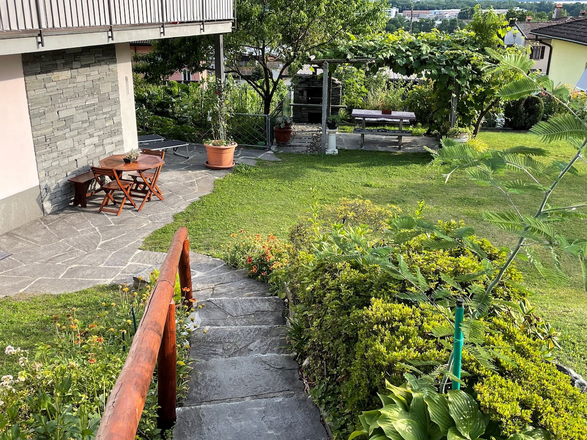 A stone pathway leads through a well-maintained garden, highlighted by greenery and blooming flowers. A wooden table with chairs is positioned on a patio, surrounded by established plants. A shaded area with a bench can be seen in the background, offering a serene outdoor setting.