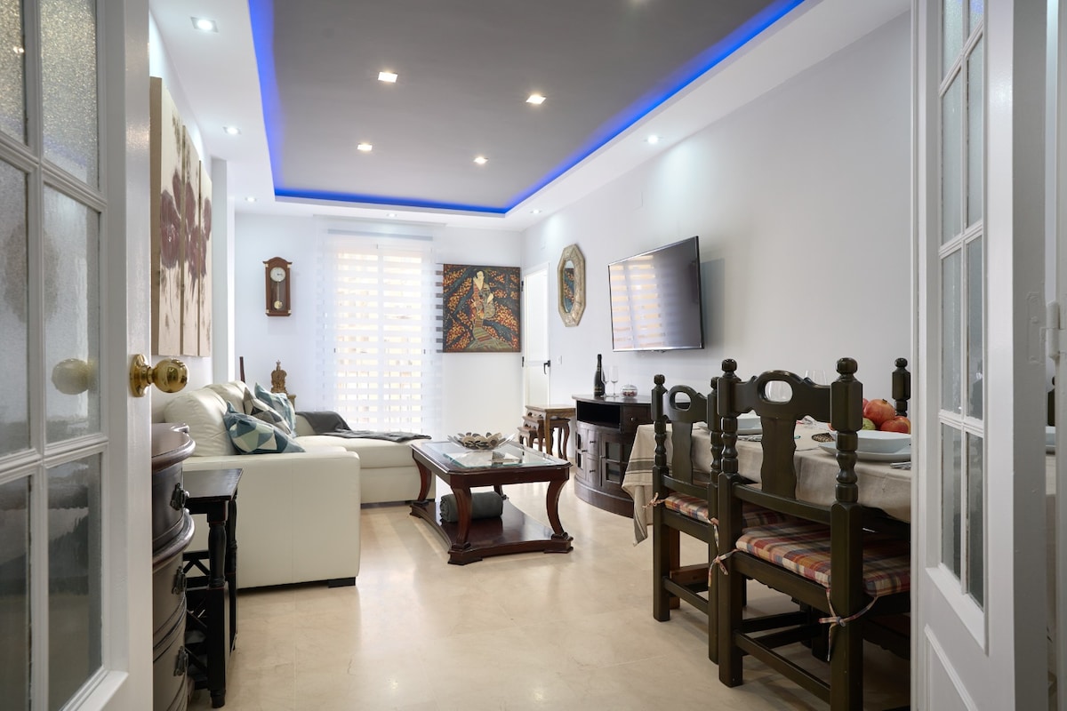 A spacious living area is featured, with modern lighting enhancing the design. A comfortable white sofa is complemented by a wooden dining table and chairs. A flat-screen television is mounted on the wall, and artwork adorns the space, contributing to a welcoming atmosphere.