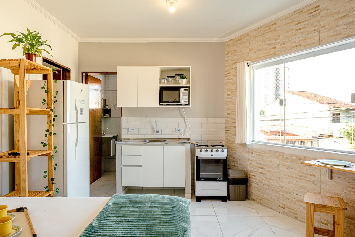 The modern kitchen area features a stove, refrigerator, and microwave, all positioned against a tiled wall. Natural light flows through a large window, illuminating the space. A wooden dining table and shelves with plants add to the minimalist design, contributing to a cozy atmosphere.