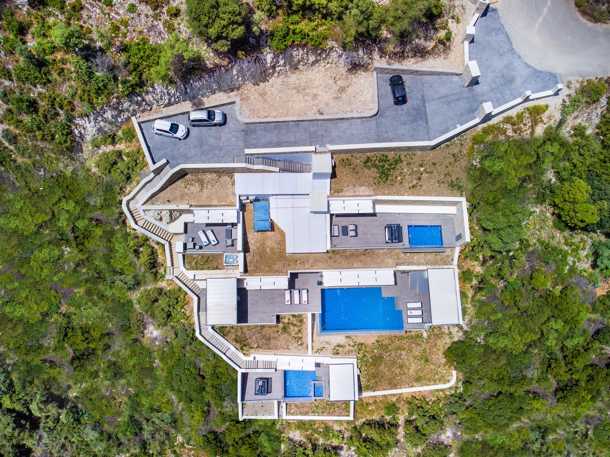 An aerial view showcases the layout of De Sun Suites – Villa Grey, highlighting multiple outdoor spaces. Private pools and sun loungers are arranged amidst lush greenery, with the accommodation and parking areas clearly visible. The surroundings reflect a serene, secluded mountain locale.