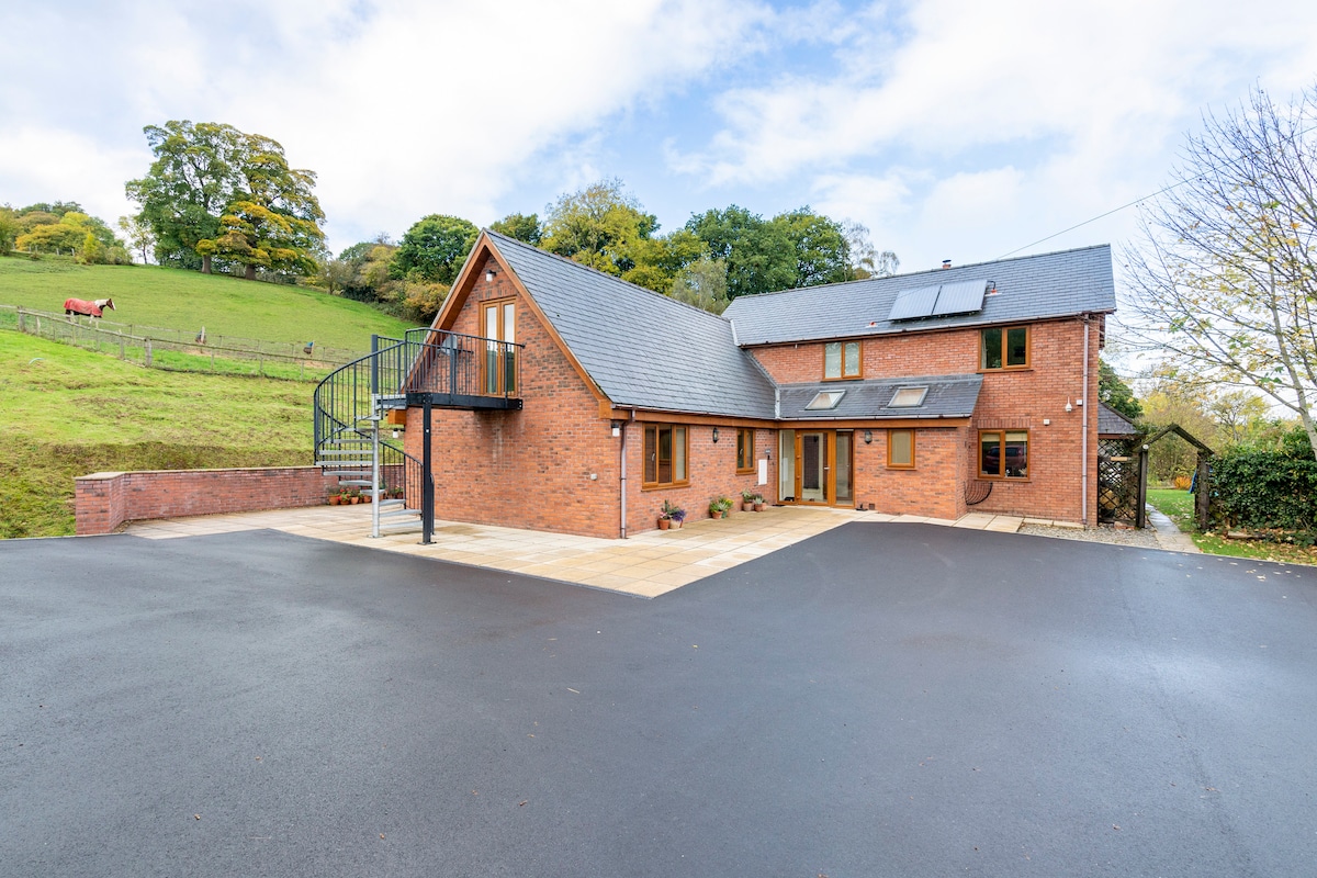 The modern brick exterior of the annexe is showcased, featuring a mix of large windows and a pitched roof. A spacious parking area is visible in front, with a backdrop of a green hillside and horses grazing in the distance. A spiral staircase leads to an upper level.