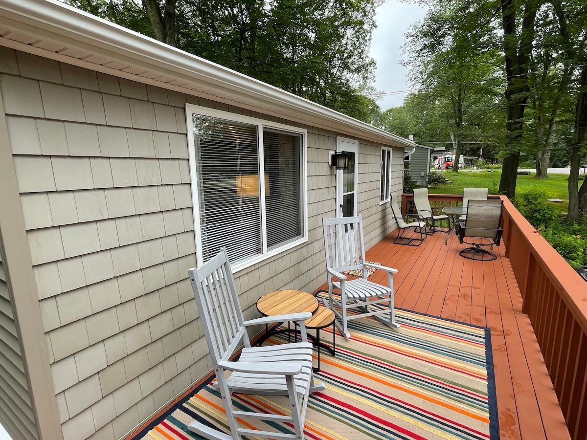 A well-maintained deck features two white chairs and a small round table. A colorful striped rug enhances the outdoor space, while trees provide shade in the surrounding area. Natural light filters through the windows of the adjacent cottage.