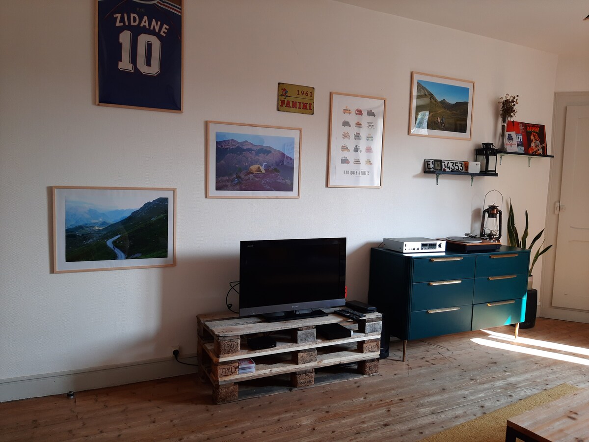 A living area is depicted with a television on a wooden pallet stand. The wall features framed artwork, including landscape photographs and decorative items. A plant adds a touch of greenery, while a console table holds additional decor. A sports jersey is displayed above.
